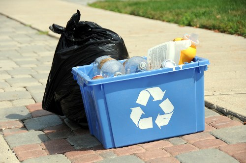 On a paved sidewalk in a residential area, a blue recycling bin with a white recycling symbol on the front is filled with clear plastic bottles, some with labels still attached, and a few other recyclable containers. To the left of the bin, a large black garbage bag is tied at the top and appears full, leaning slightly against the bin. The background includes a strip of green grass and part of a concrete curb, with a grassy verge beyond. The scene is well-lit, suggesting daylight, and the setting indicates rubbish collection or removal activity typical of local waste management services in Ickenham, as provided by Commercial Waste Removal Ickenham, supporting waste disposal needs in the area.