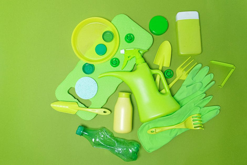 A collection of various cleaning tools and supplies arranged on a flat surface, including a bright green plastic watering can, a pair of matching rubber gloves, a small yellow-handled trowel, a green 