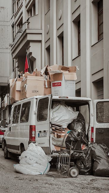 A white commercial van with its rear doors open, parked on a city street in front of tall, beige-colored buildings. The van's cargo area is filled with various types of rubbish, including large black