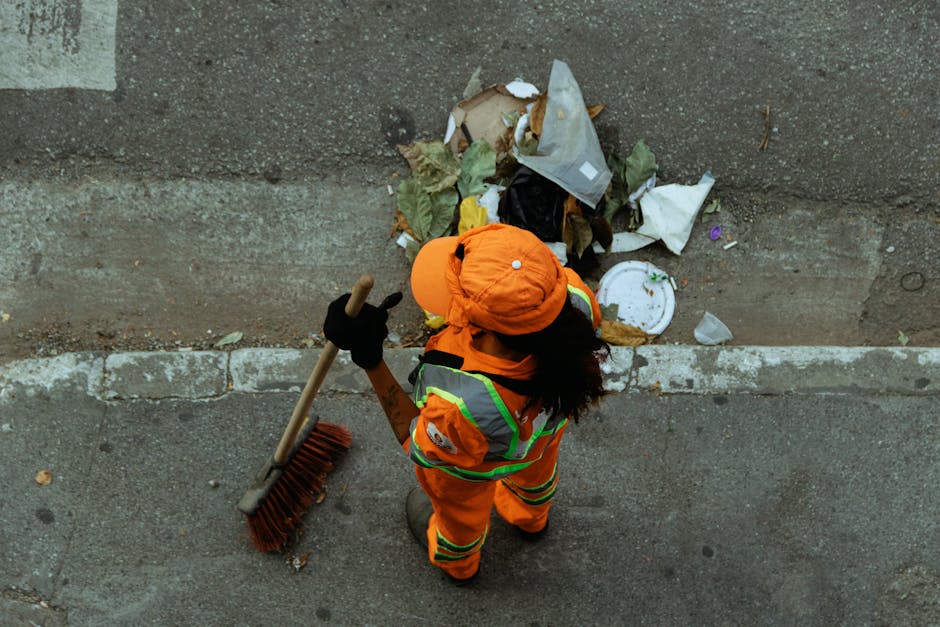A waste collection operative, dressed in a high-visibility yellow and red vest, is seen loading rubbish into the rear compartment of a large red refuse collection vehicle parked on the side of a stree